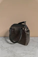 کیف دستی زنانه چرم قهوه‌ای تیره با طرح پوست ماری و دسته‌های مشکی، نمای سه‌رخ از سمت راست روی سطح مرمر با پس‌زمینه بژ روشن
