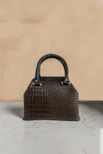 کیف دستی چرم زنانه قهوه‌ای تیره با طرح پوست ماری و دسته‌های مشکی، قرار گرفته روی سطح مرمر روشن با پس‌زمینه‌ی بژ